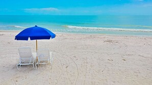 Ein Strand mit einem blauen Sonnenschirm und zwei weißen Liegestühlen.