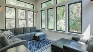 A modern living room with a sectional sofa, ottoman, and a small table. Large windows provide a view of the outdoors. - Ski & Hike in Kelly Canyon: Rigby Group Getaway (Rigby)