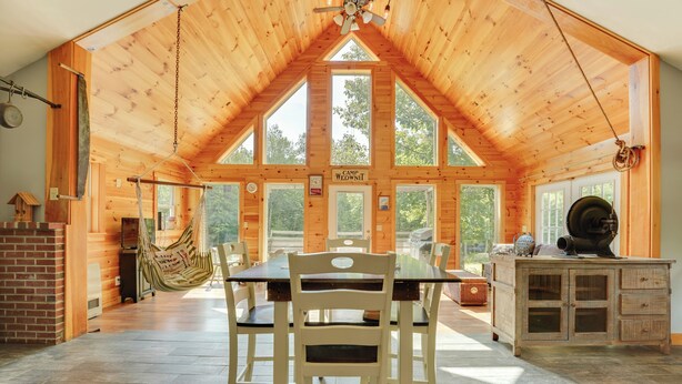 A spacious wooden cabin with a high ceiling, large windows, a dining table, chairs, a hammock, and a fireplace. - Fish + Hike: Wooded Home w/ Deck in Dexter (Dexter)
