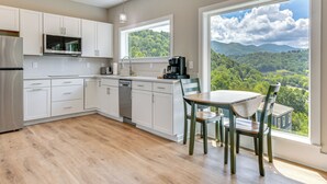 A modern kitchen with white cabinets, a stainless steel refrigerator, a dishwasher, and a coffee maker. There is a dining table with chairs and a large window offering a view of a forested landscape. - Smoky Mtn Getaway: Modern 2br/2ba W/stunning Views (Maggie Valley)
