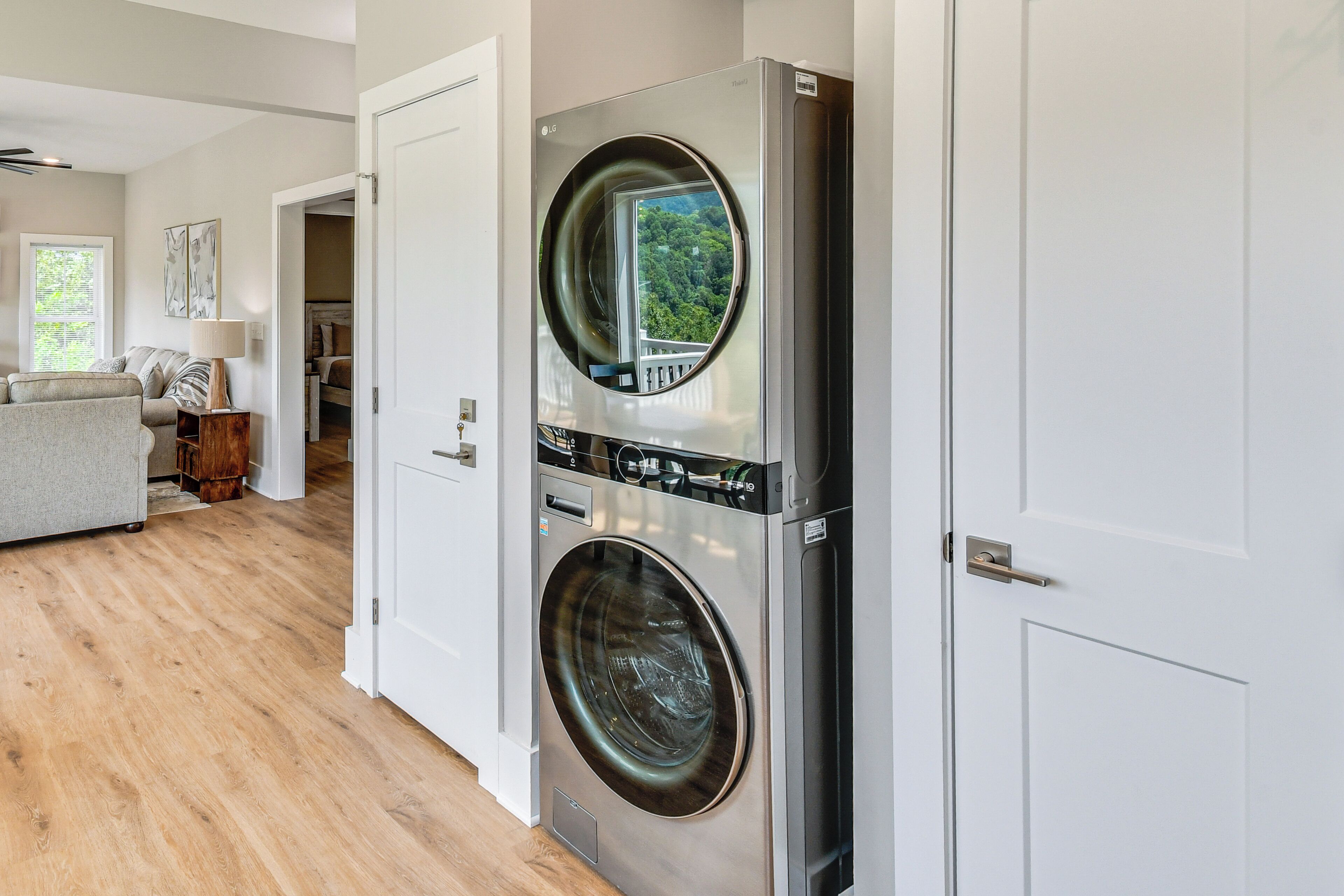 A laundry room with two stacked front-loading washing machines, a wooden floor, and a view of a green landscape through the door.