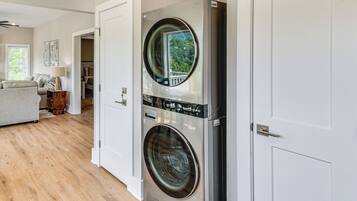 A laundry room with two stacked front-loading washing machines, a wooden floor, and a view of a green landscape through the door.