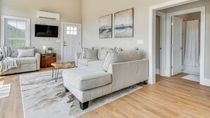 A spacious living room with a large grey sectional sofa, a coffee table, and a flat-screen TV mounted on the wall. There is a wooden floor and a large window with blinds. - Cozy 2br/2ba Mtn Escape w/ Views in Maggie Valley (Maggie Valley)