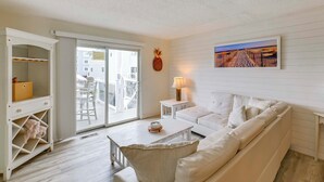 Een woonkamer in witte tinten, met een bank, salontafel en lamp. Er is een glazen schuifdeur die leidt naar een buitenruimte met stoelen.