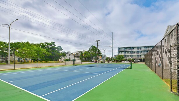 Een tennisbaan buiten met een groene omgeving, een hek en een gebouw op de achtergrond.