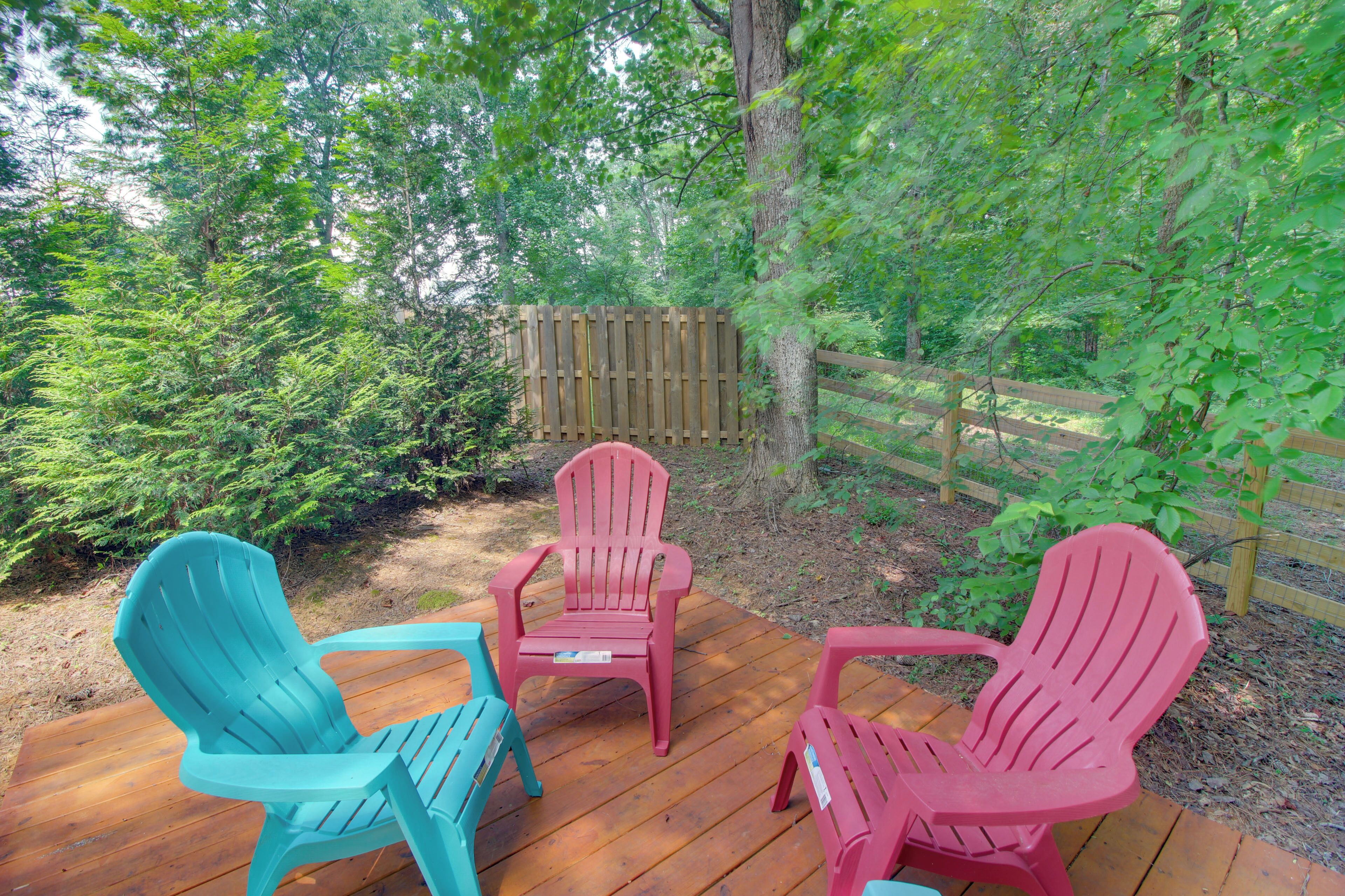 Une terrasse en bois avec trois chaises Adirondack, entourée d’une végétation luxuriante et d’une clôture en bois.