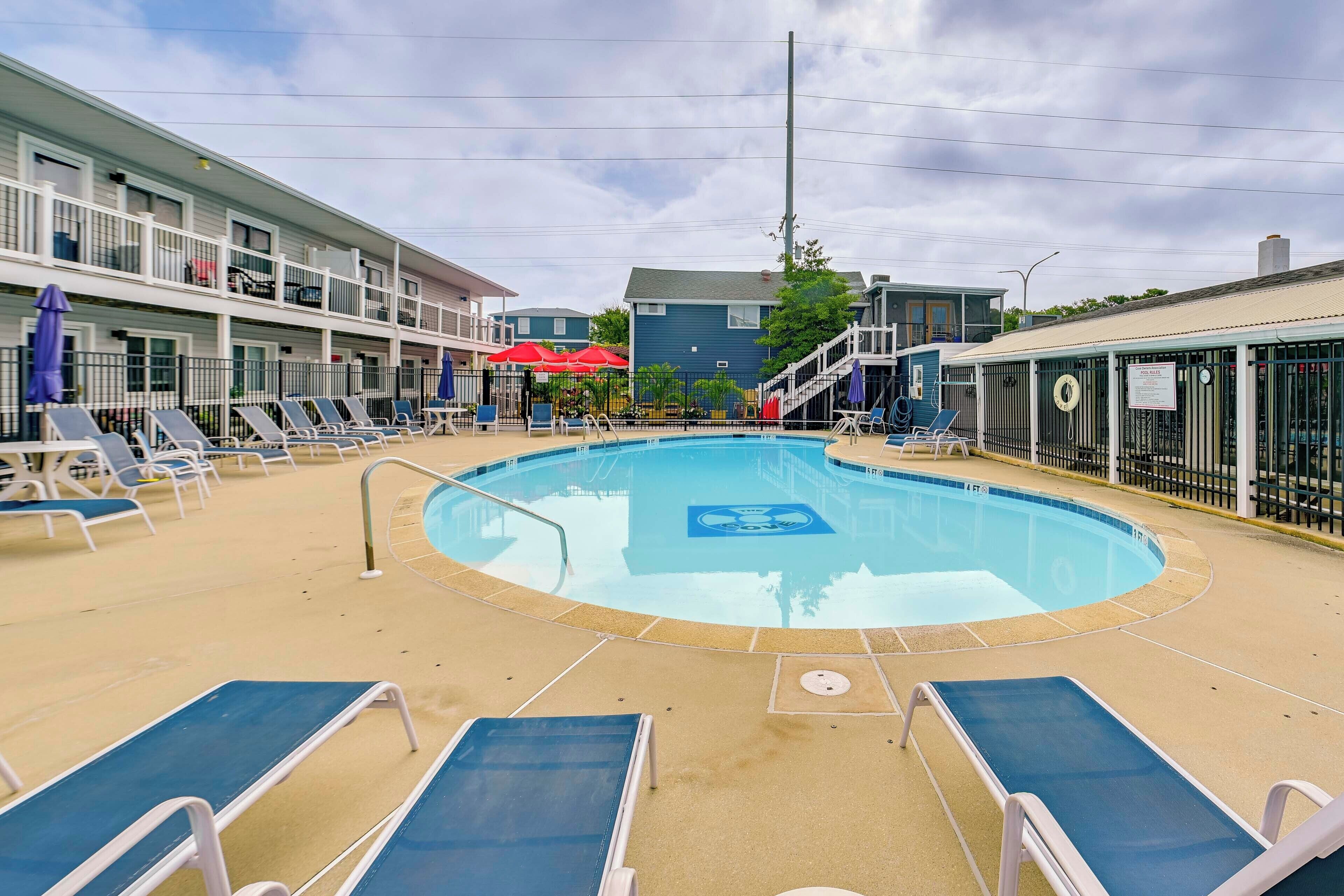 A pool area with lounge chairs and umbrellas, a pool with a lifeguard stand, and a building with balconies.