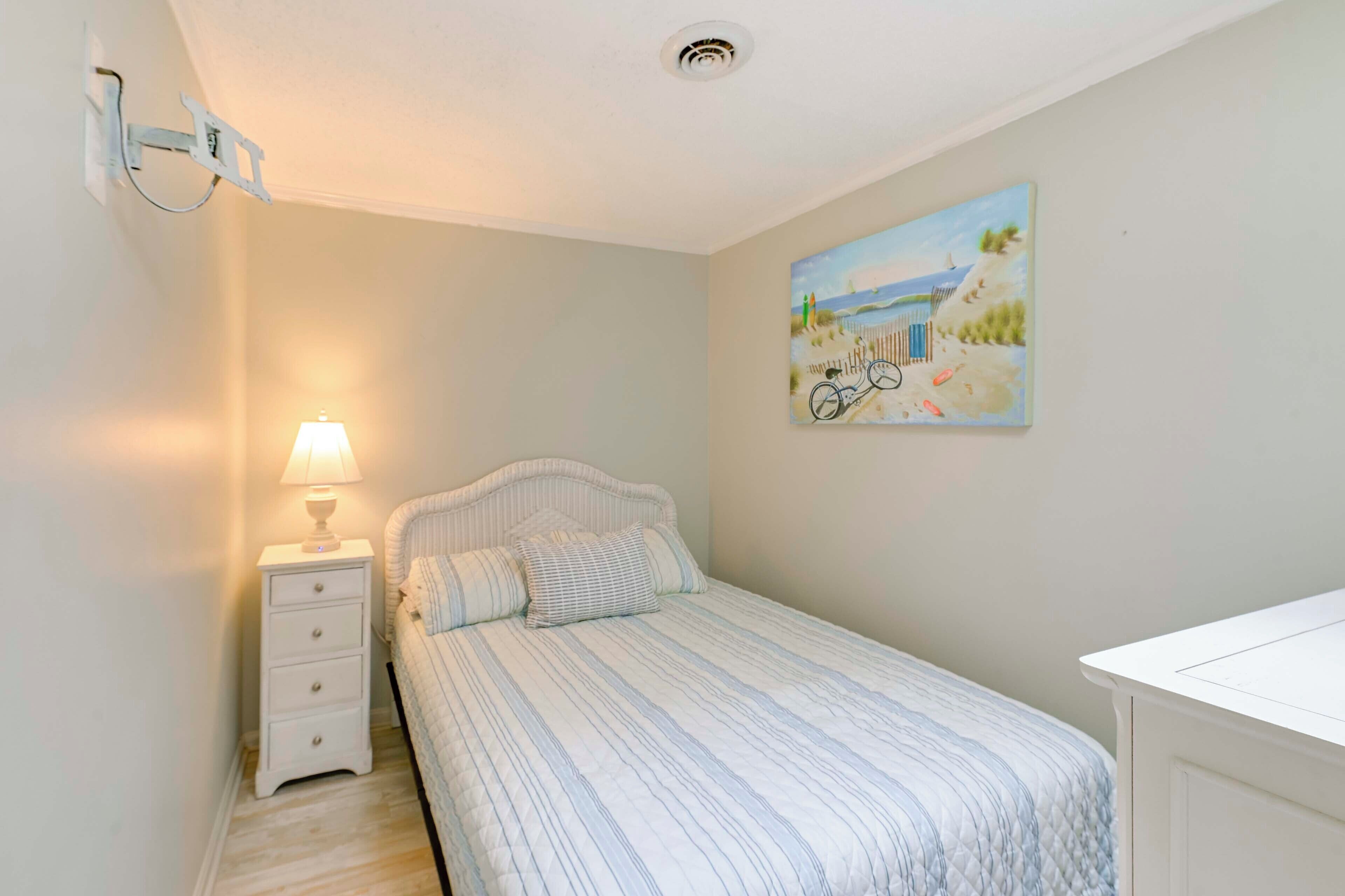 A bedroom with a bed, a nightstand with a lamp, a wall-mounted light fixture, and a beach-themed painting.