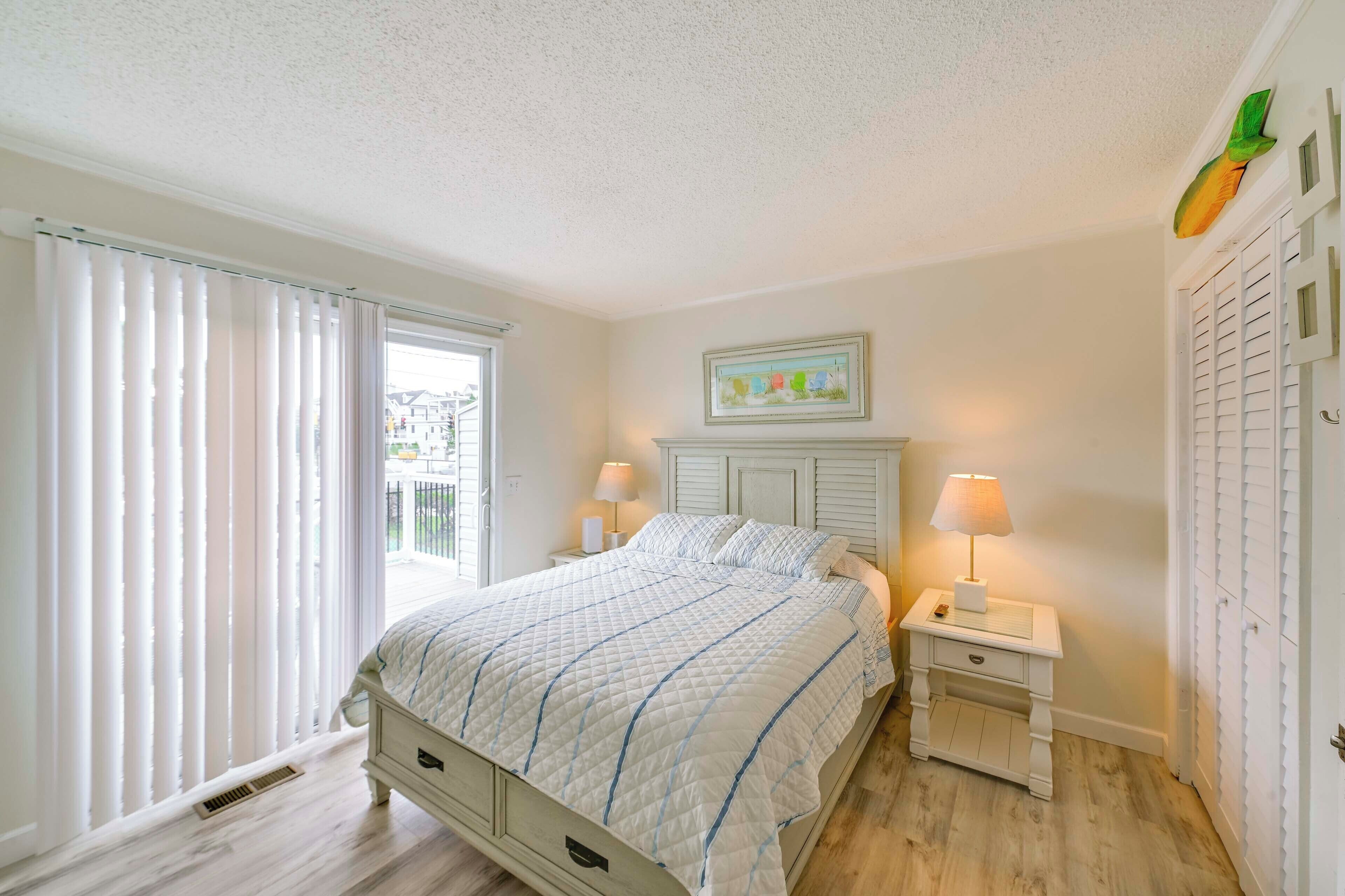 Un dormitorio con cama, dos mesitas de noche con lámparas, una ventana con cortinas, una puerta decorada con motivos de frutas y un cuadro en la pared.