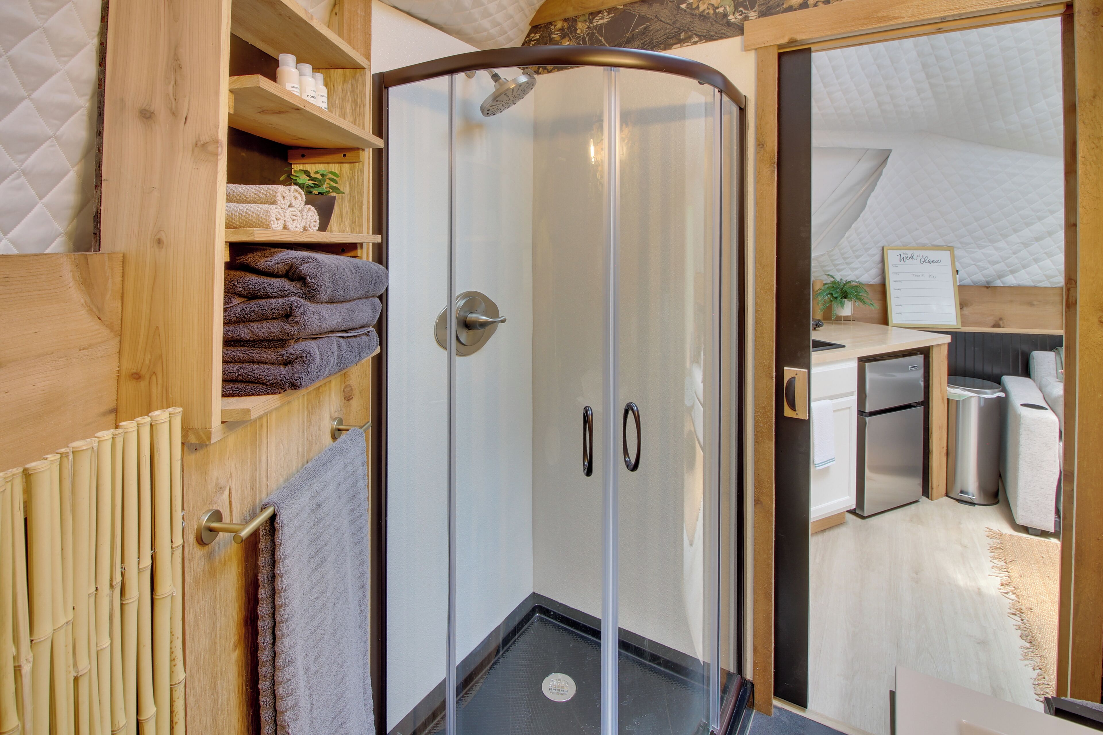 A modern bathroom with a glass shower enclosure, wooden shelves with towels, and a visible kitchen area.