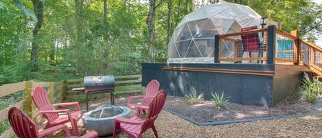 Une structure en forme de dôme géodésique, dotée d’une terrasse en bois et d’un espace salon, le tout entouré d’un environnement boisé.