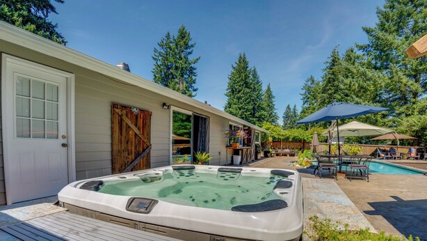 A hot tub in a backyard with a wooden deck, a pool, and a covered patio area with outdoor furniture.