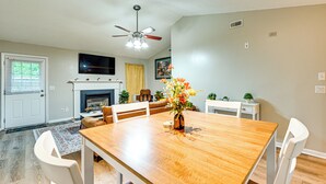 A dining area with a wooden table, white chairs, a vase with flowers, a ceiling fan, and a fireplace.