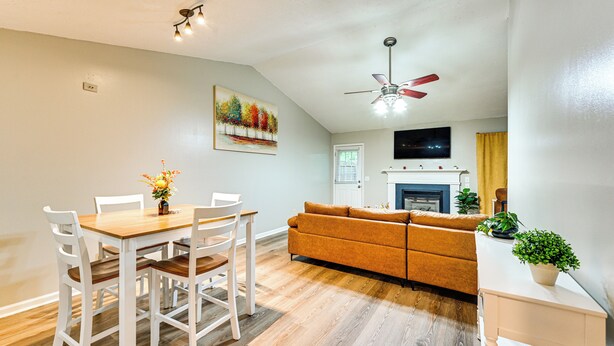 A modern living room with a wooden dining table, white chairs, a sofa, and a ceiling fan.