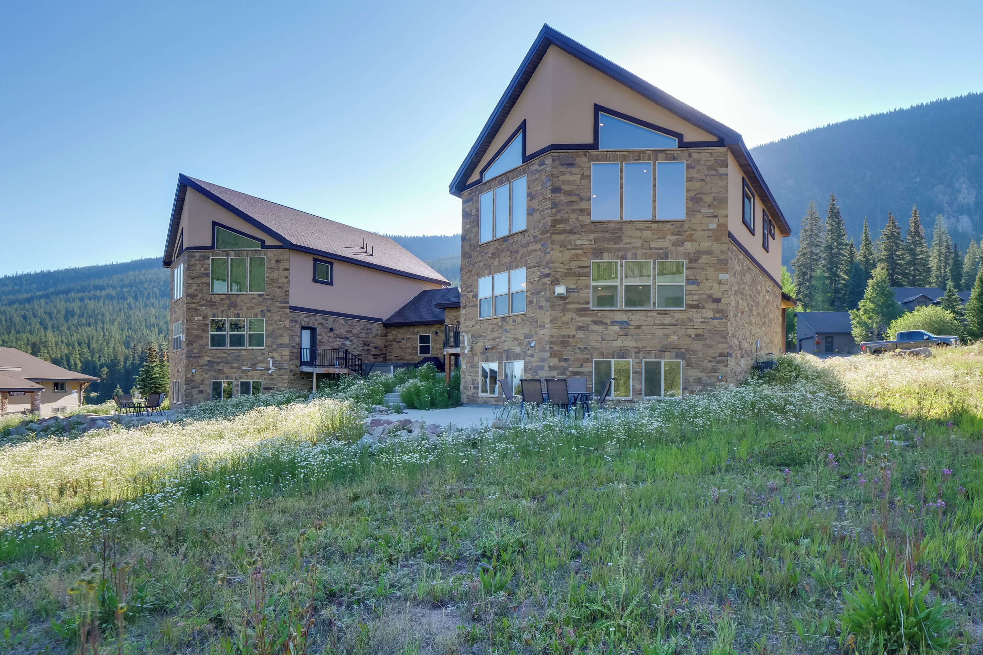 A modern two-story house with a stone facade, large windows, and a sloped roof, surrounded by a grassy area and mountains in the background.