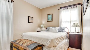 Un dormitorio con una cama grande, un cofre de madera, dos lámparas de noche y una ventana con cortinas.