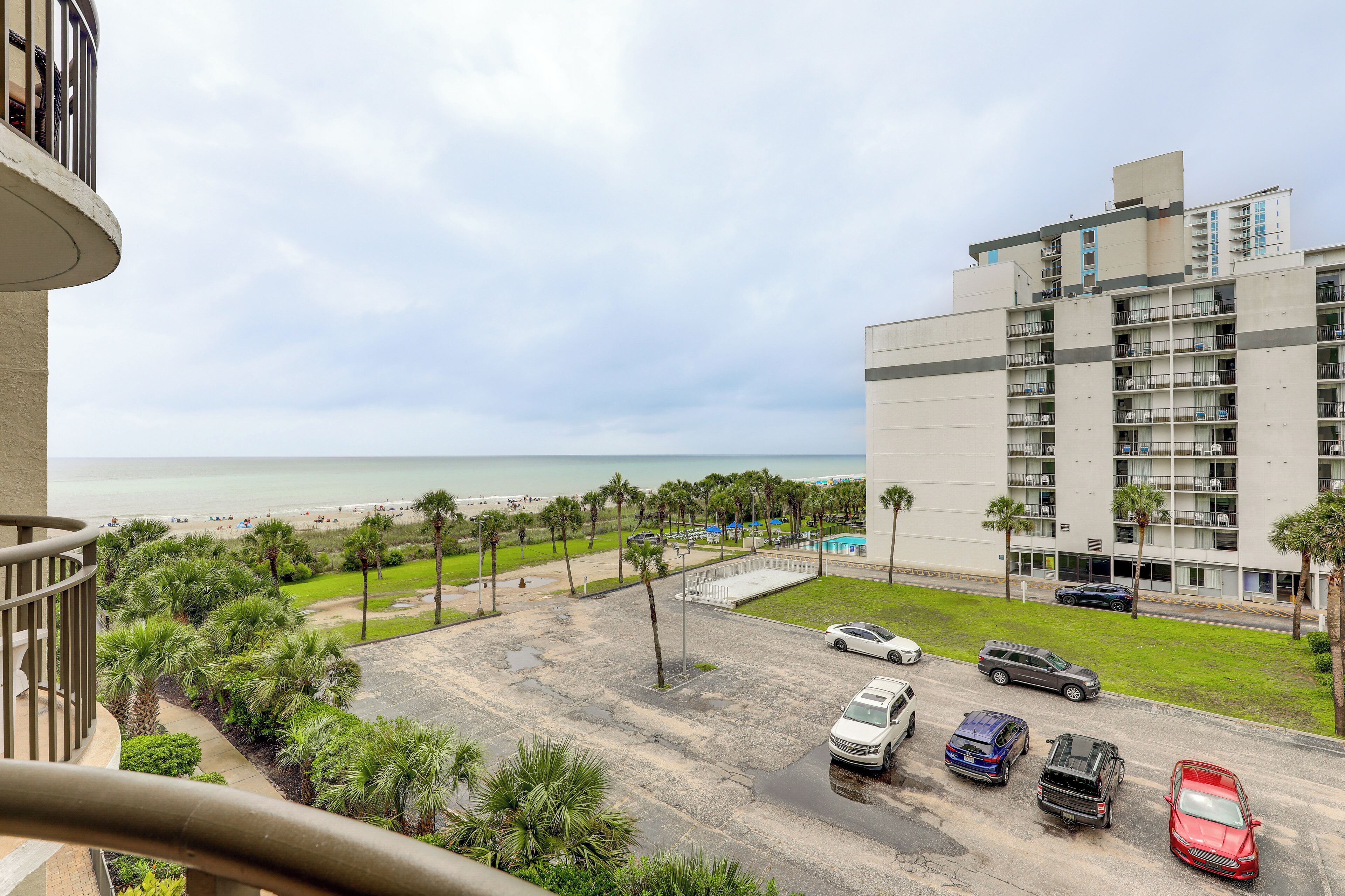 A view from a balcony overlooking a parking lot with several cars, a beach, and a hotel building with multiple floors and balconies.