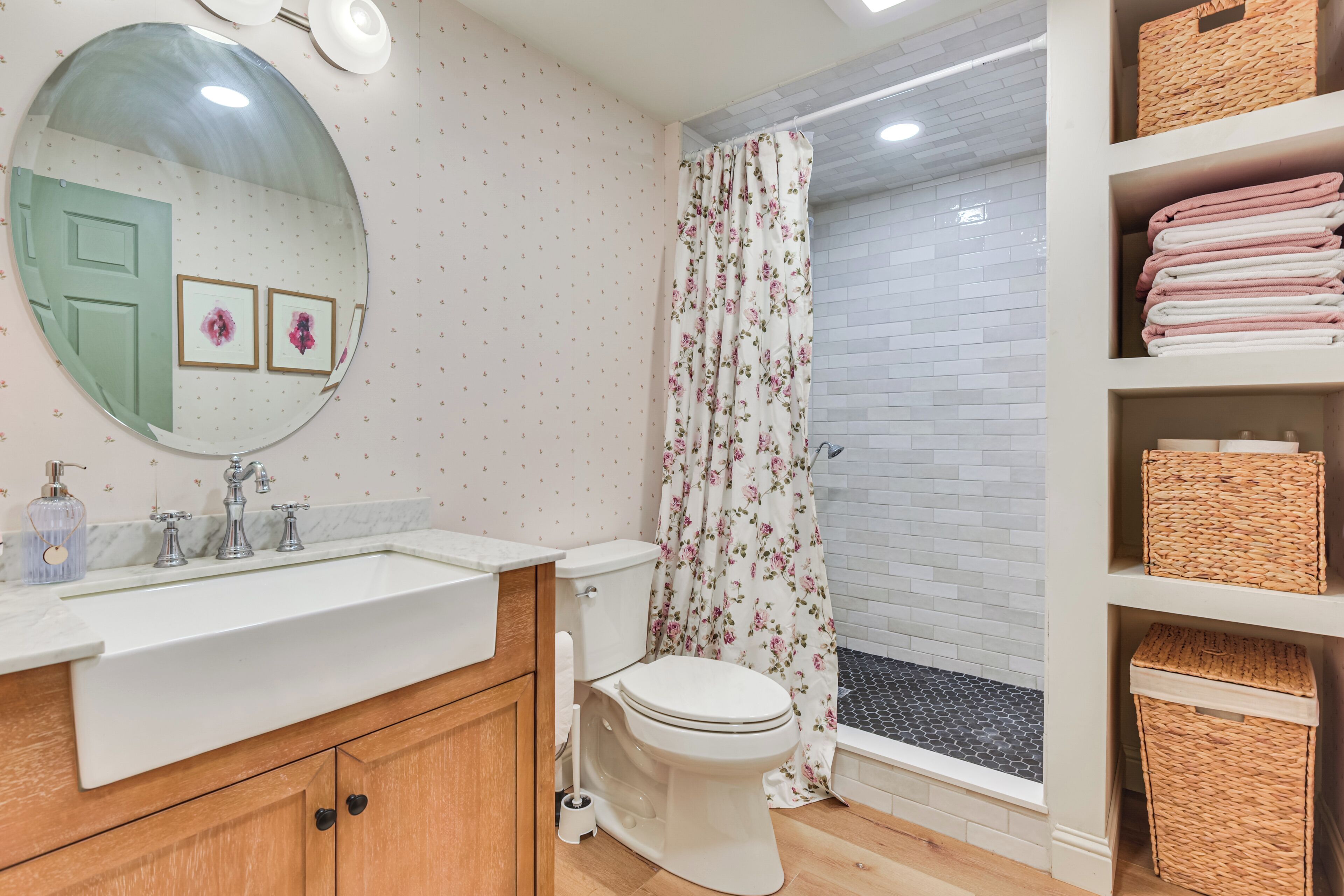 Une salle de bain avec un lavabo blanc, des meubles en bois, des toilettes, une douche avec un rideau à motifs floraux et un carrelage au sol.