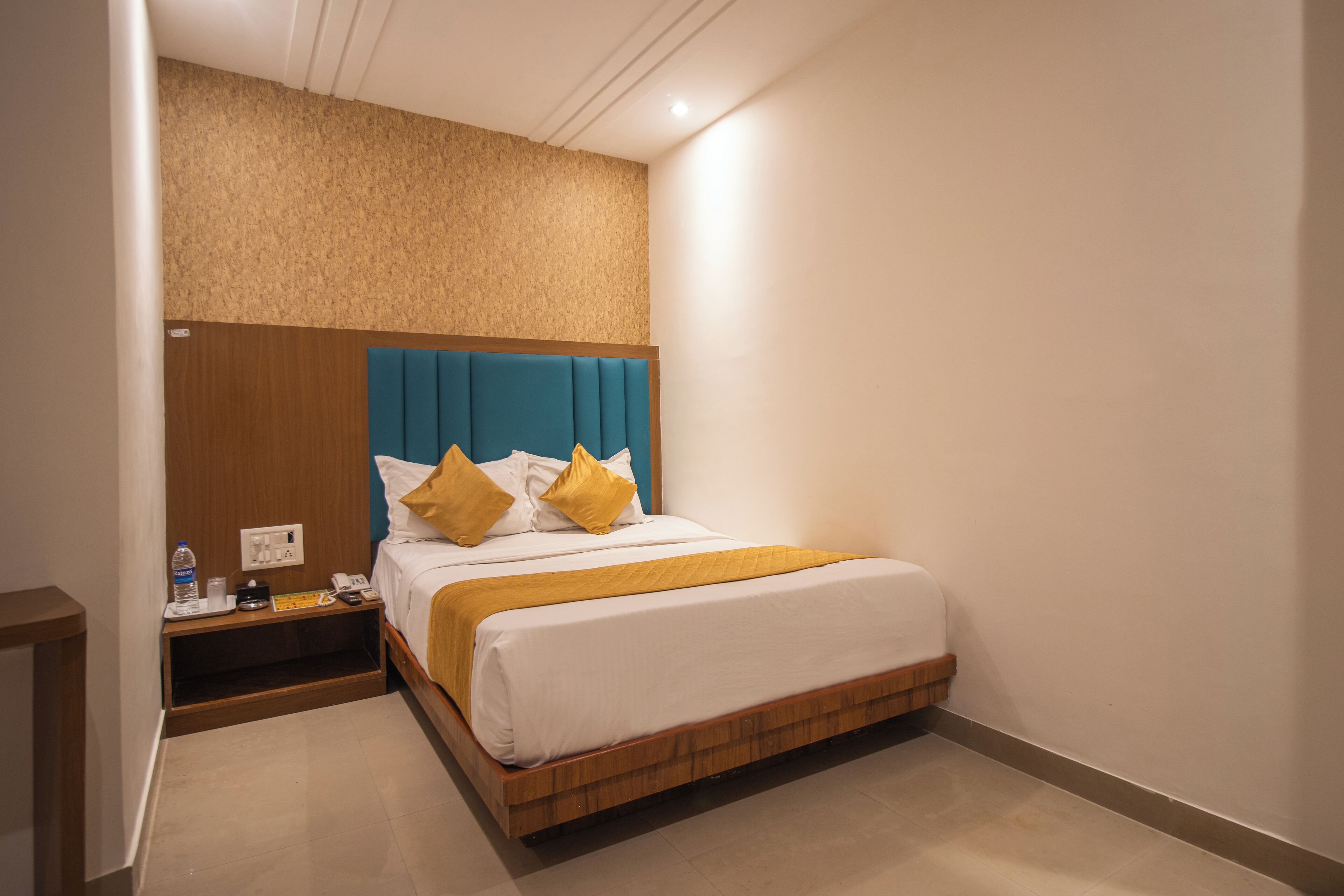 A hotel room with a wooden headboard, a bed with white and yellow bedding, a nightstand with a bottle and phone, and a wall-mounted shelf.