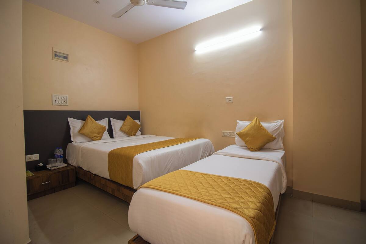 Habitación de hotel con dos camas individuales, cada una con ropa blanca y un plaid amarillo. Hay una mesita de noche con una lámpara y una botella de agua. Las paredes son color beige y tiene un ventilador de techo.