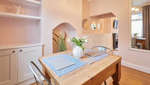 A dining area with a wooden table, blue-striped placemats, and a white vase with flowers. There are white cabinets and a mirror on the wall. - Host Stay Horner Street (York)