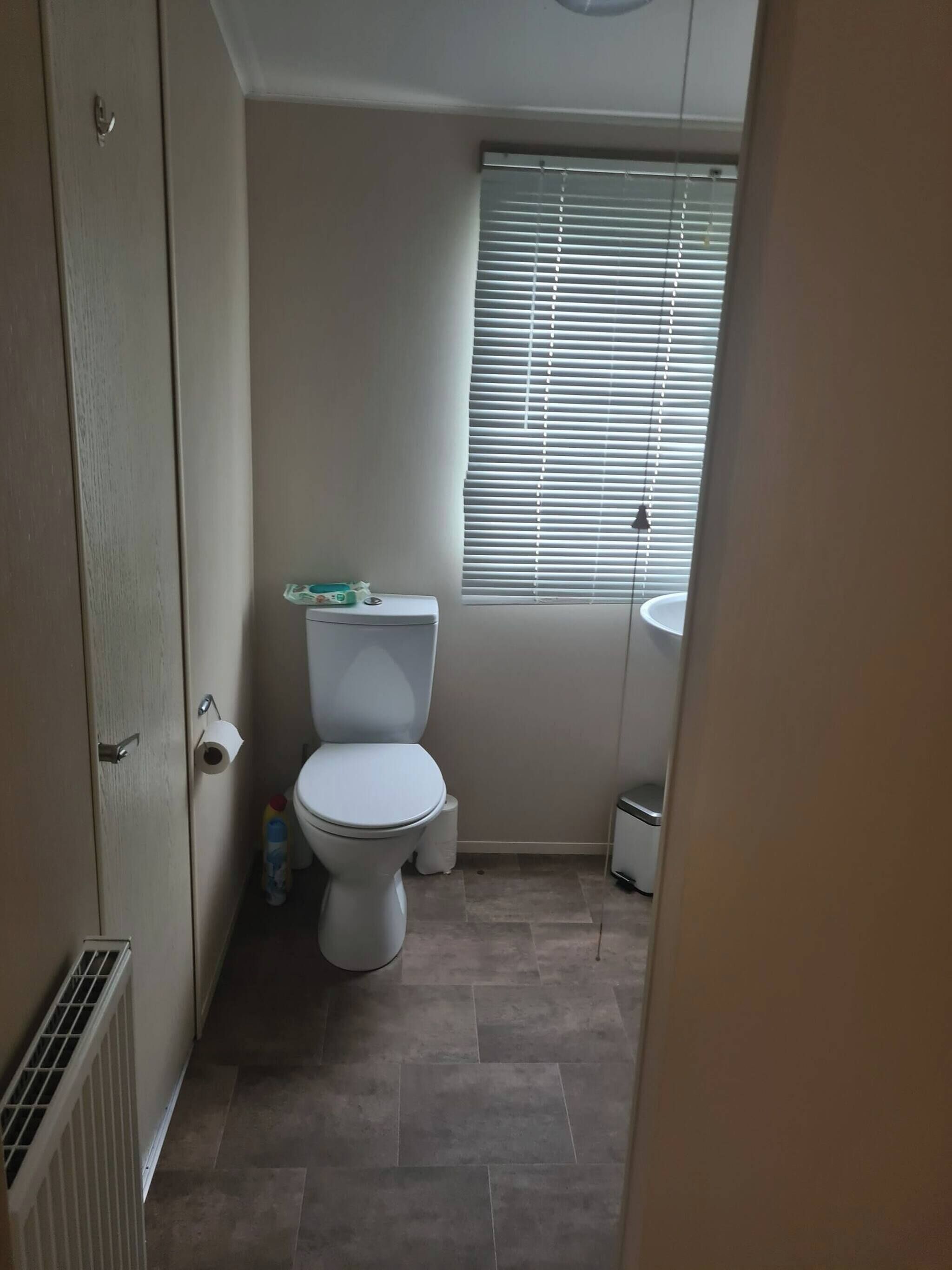 A small, clean bathroom with a toilet, a window with blinds, a sink, and a toilet paper holder.