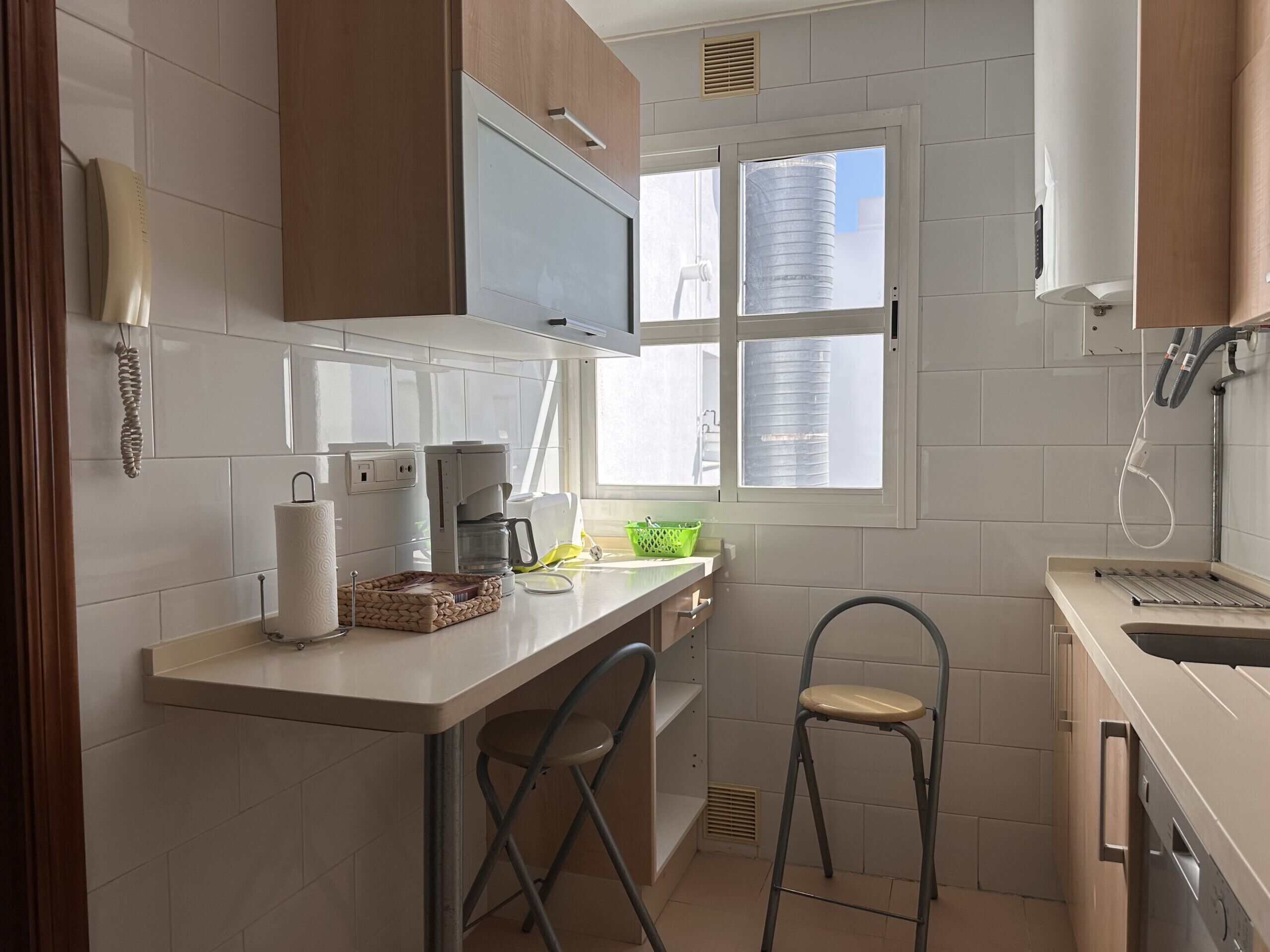 Een kleine keuken met witte tegels, een raam met zonwering, een koffiezetapparaat en een spoelbak.