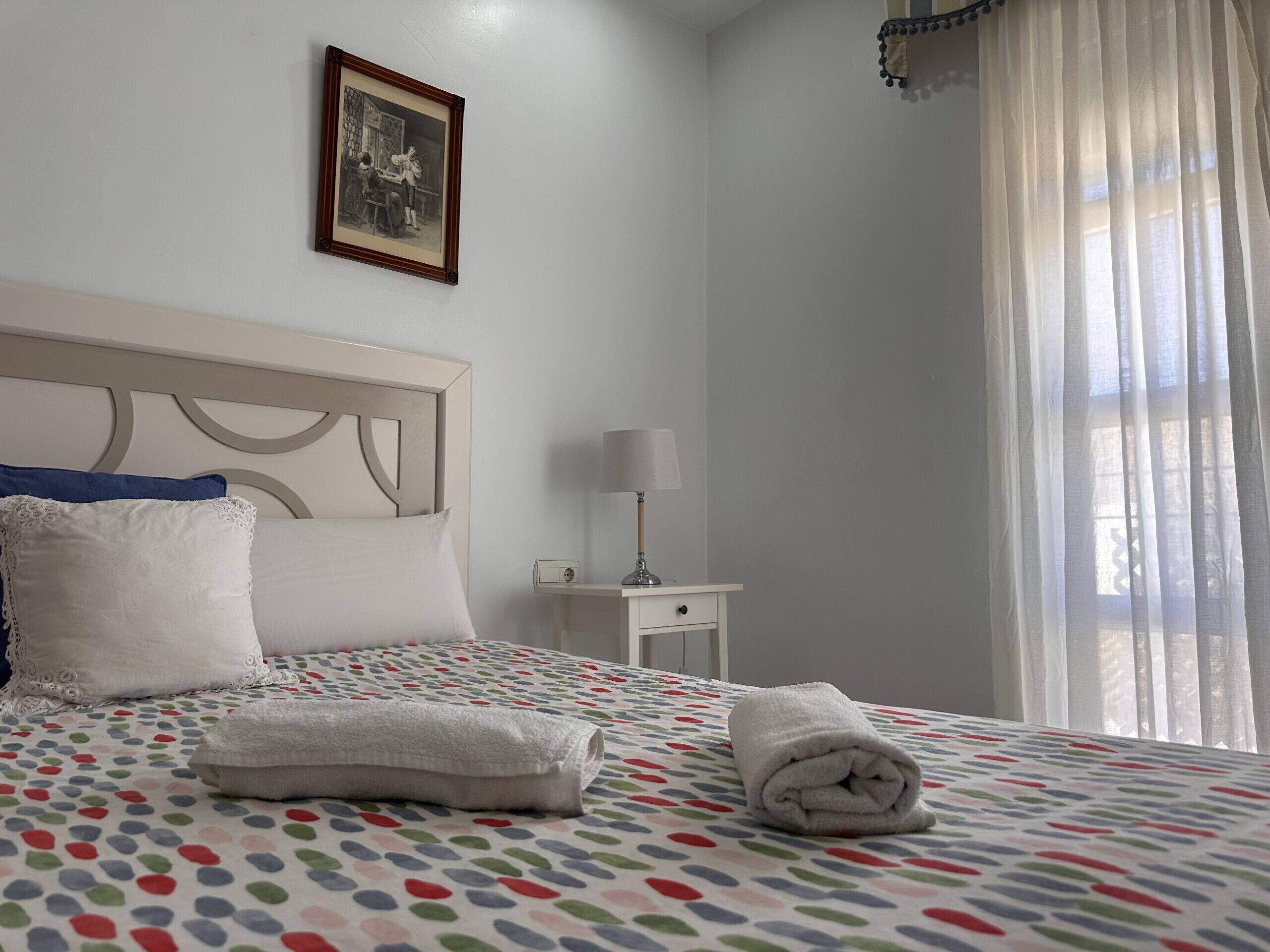 Een net opgemaakt bed met een stippenbed overtrek, een wit kussen en een opgerolde handdoek aan de zijkant. Er staat een klein nachtkastje en erboven een fotolijst aan de muur.