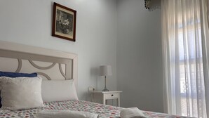 A neatly made bed with a polka dot bedspread, a white pillow, and a rolled towel on the side. There is a small bedside table with a lamp and a framed picture on the wall above the bed.