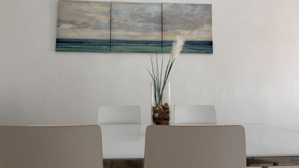 A dining area with a table and chairs, a painting of a seascape, and a vase with dried plants.
