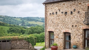 A stone building with a rustic facade, a large potted plant in front, and a view of rolling hills in the background.