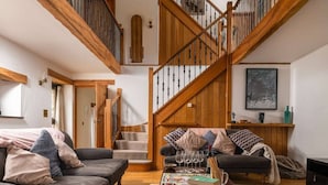 A spacious living room with a wooden staircase, a dark-colored sofa, and a coffee table with books and glasses.