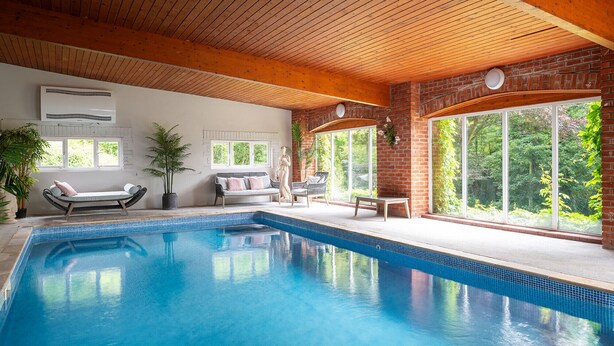 Una piscina interior con techo de madera, pared de ladrillo y grandes ventanales que ofrecen vistas al verdor.
