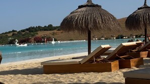 A beach with wooden lounge chairs and thatched umbrellas, a clear blue sky, and a swan-shaped boat in the water.