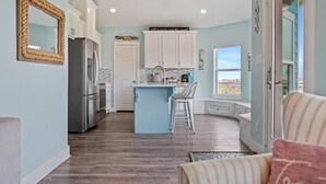 A modern kitchen with white cabinets, a central island, and a view to the outside. - Steps to the Beach | Tiki Bar Fun | Epic Bay Views | Sleeps 8 | Pet-Friendly (Crystal Beach)
