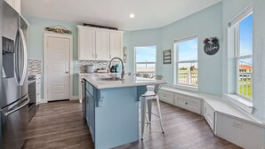 A modern kitchen with a central island, white cabinetry, and a view of a fenced yard through large windows. - Steps to the Beach | Tiki Bar Fun | Epic Bay Views | Sleeps 8 | Pet-Friendly (Crystal Beach)
