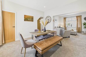 A modern living room with a wooden dining table, cushioned seating, and a large window with curtains. - Host & Stay | Jomor House (Burnhope)