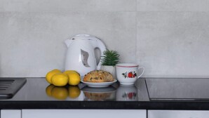 A kitchen with white cabinets, a black countertop, a white kettle, lemons, a plate with a pastry, a small potted plant, and a white mug with red trim.