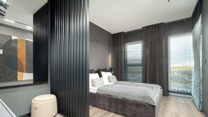 A modern bedroom with a bed, a round ottoman, a large window with blinds, and a partial view of a bathroom.