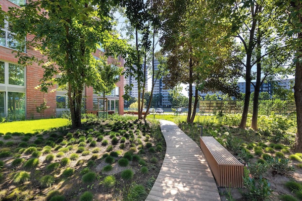 A pathway through a landscaped area with trees and shrubs, leading to a building with large windows and a brick facade.