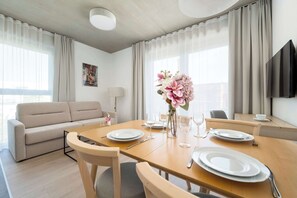 Ein modernes Wohnzimmer mit Sofa, Esstisch für zwei Personen, Blumenvase und Wand-Fernseher.