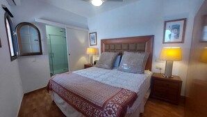 A bedroom with a bed, two bedside lamps, a wooden headboard, a mirror, and a door leading to a bathroom. - Apartment in the centre of Mijas Pueblo with a large terrace and sea views (Mijas)