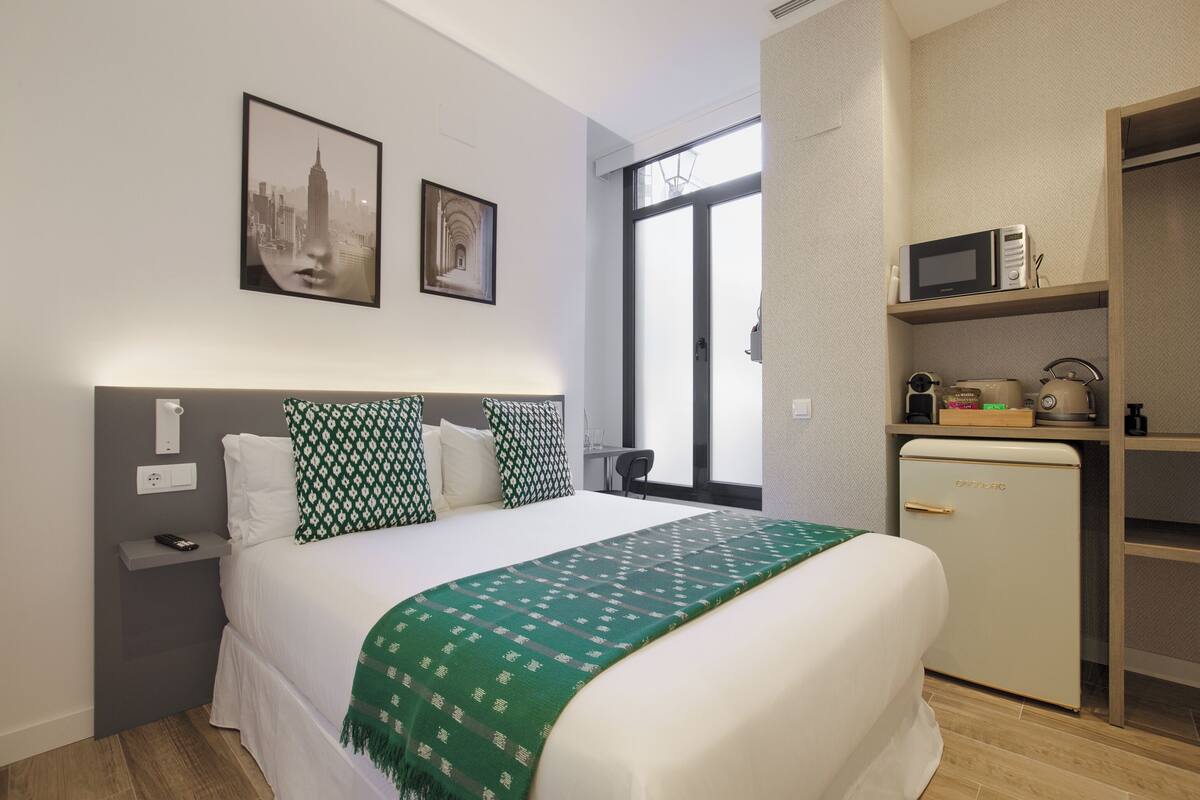 A neatly arranged bedroom with a bed, a nightstand, a dresser, a microwave, and a window with blinds.