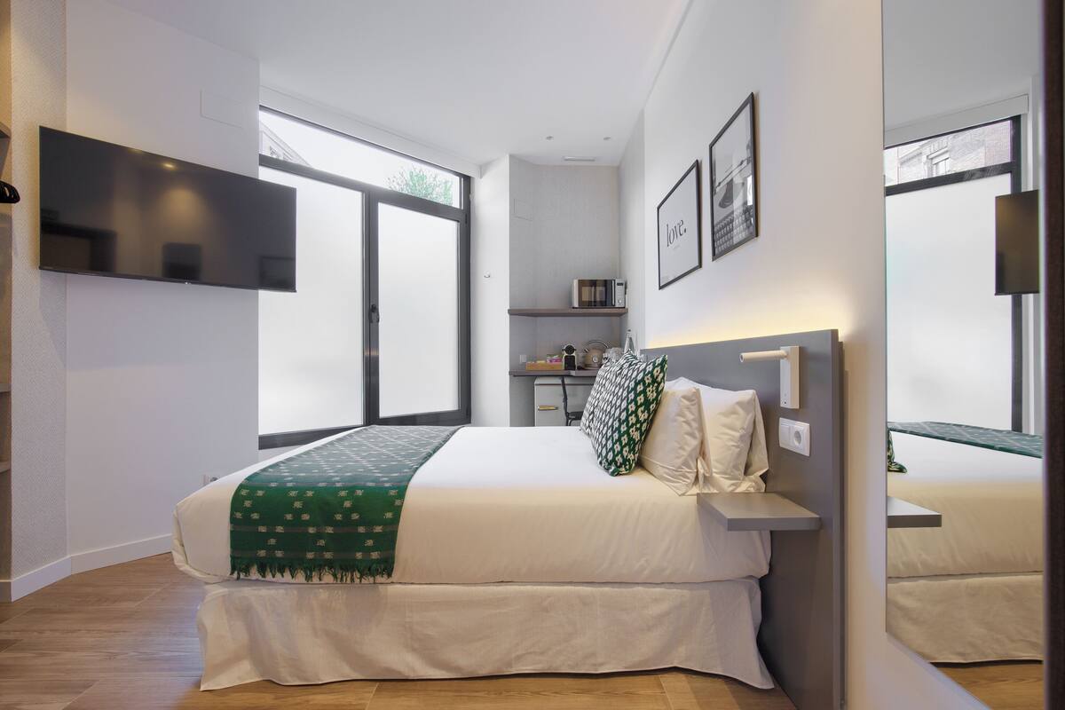 A modern bedroom with a large bed, a flat-screen TV mounted on the wall, a bedside table, and a window with a view.