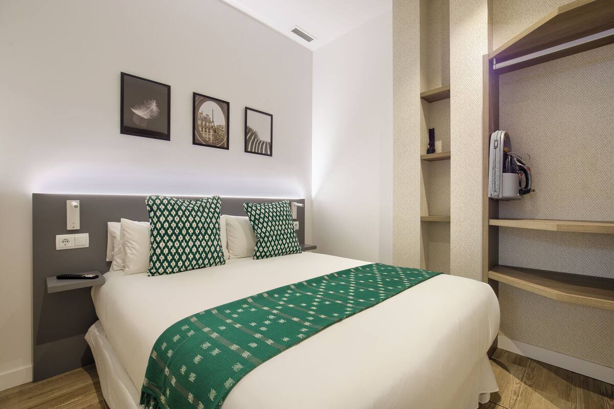 A neatly arranged hotel room with a bed, a bedside table, and a shelf with a suitcase.