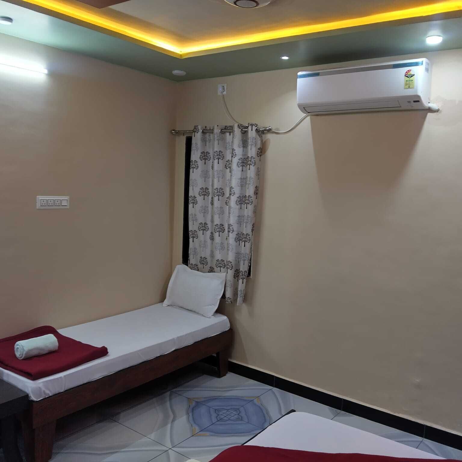 A small, single-bed room with a wooden bed frame, a white mattress, a red blanket, and a patterned curtain. An air conditioning unit is mounted on the wall.