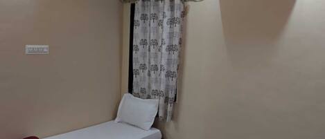 A small, single-bed room with a wooden bed frame, a white mattress, a red blanket, and a patterned curtain. An air conditioning unit is mounted on the wall.
