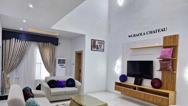 A modern living room with a large TV, comfortable seating, and a sign reading "Wuraola Chateau". - DFW Abodes Luxury Home (Akure)