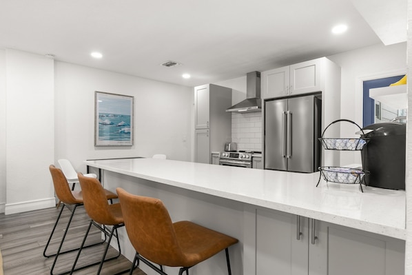 Una cocina moderna con armarios blancos, electrodomésticos de acero inoxidable y encimera blanca. Hay tres sillas marrones alrededor de la barra.