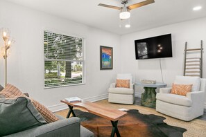A living room with a gray sofa, two white armchairs, a wooden coffee table, a television mounted on the wall, and a ceiling fan.